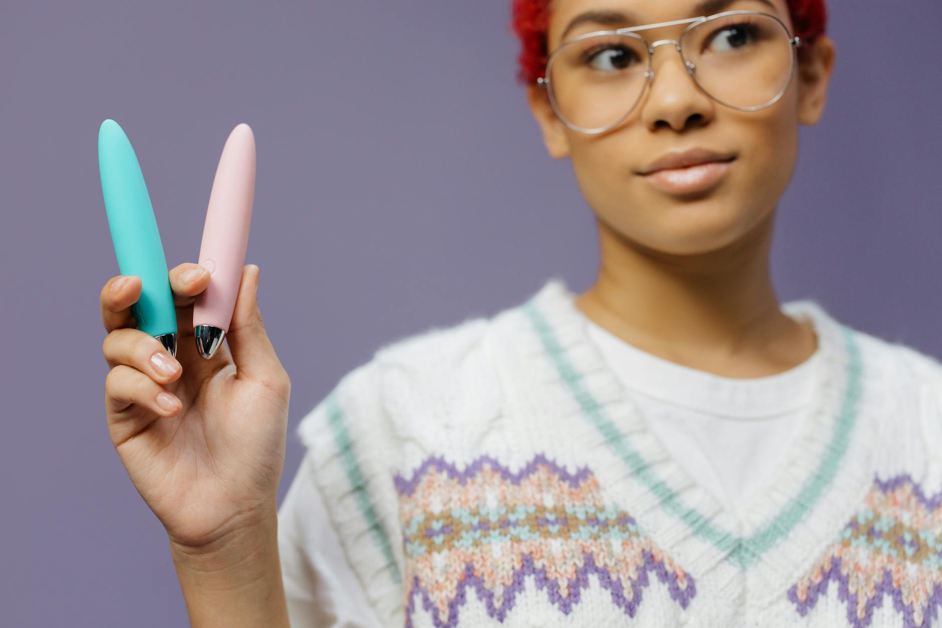 A hand holding two colorful clitoral vibrators against a purple background